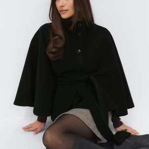 Svart poncho kappa med bälte och hög krage - Stilren svart poncho kappa med vid siluett och hög krage. Jackan har knappar framtill och ett knytbälte i midjan för en snygg passform. Ärmarna är vida och ger en cool cape-känsla. Perfekt för dig som vill ha en klassisk men trendig look.