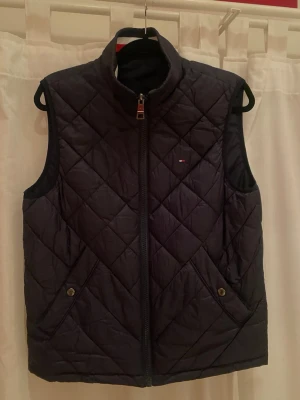 Mörkblå quiltad dunväst Tommy Hilfiger - Säljer en mörkblå quiltad dunväst från Tommy Hilfiger. Västen har dragkedja framtill, två fickor med knapp och en liten logga på bröstet. Ena sidan är helblå med quiltat mönster, andra sidan har en sportig krage med röd och vit rand. Det går alltså bra att använda den på båda sidorna! 