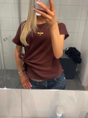 Brun t-shirt med knyt i sidan - Säljer en brun t-shirt med korta ärmar och snygg knytning i sidan. T-shirten har en enkel, sopren look och är gjord i mjukt bom sopmaterial. Passformen är normal och den är perfekt för dig som gillar stilrena plagg med en twist.