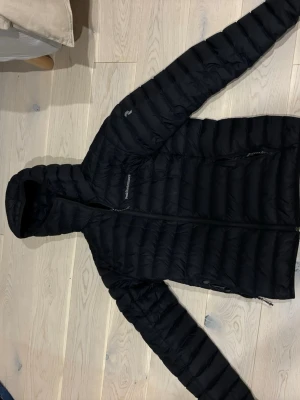 Svart dunjacka från Peak Performance - Säljer en svart dunjacka från Peak Performance med huva och dragkedja framtill. Jackan är quiltad och har en clean look med diskret logga på bröstet. Perfekt för kalla dagar och enkel att matcha med allt.