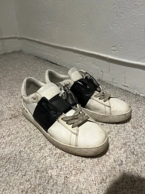 Valentino Garavani vita sneakers - Säljer ett par vita Valentino Garavani sneakers i skinn med bred svart läderdetalj över sidan och coola nitar baktill. Klassisk låg modell med snörning och rund tå. Perfekta för dig som vill ha en lyxig och edgy touch till din outfit.