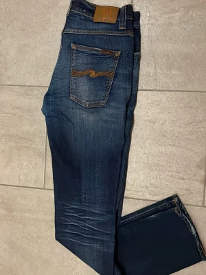 Nudie Jeans Grim Tim blå jeans 33/34 - Säljer ett par Nudie Jeans Grim Tim i tvätten Cold Crisp. Klassiska blå jeans med snygga slitningar och kontrastsömmar. Modellen har normal passform och raka ben. Tillverkade i 98% ekologisk bomull och 2% elastan för extra komfort.