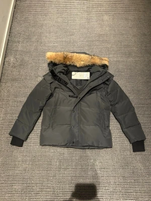 Mörkgrå dunjacka med pälskant - Säljer en mörkgrå dunjacka från Moose Knuckles med avtagbar huva och snygg pälskant i beige. Jackan har långa ärmar med muddar, två stora fickor framtill och stängs med både dragkedja och knappar. Perfekt för kalla dagar och riktigt stilren.