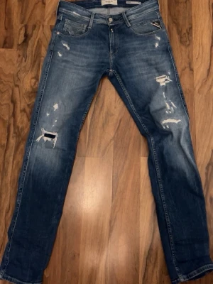 Replay jeans  - Säljer ett par blå jeans från Replay med snygga slitningar och hål på både fram- och baksida. Jeansen har klassisk femficksdesign, raka ben och normal passform. Materialet är jeans i bomull och de har en cool, sliten look. STORLEK 29/32