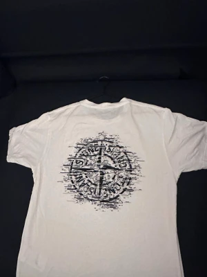 Vit Stone Island t-shirt med tryck - Säljer en clean vit t-shirt från Stone Island i bomull. Framtill finns en liten logga på bröstet och på ryggen ett stort grafiskt tryck med Stone Island-kompass. Klassisk passform och rund hals.    Äkta! 