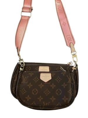 Väska - Säljer en axelväska i brunt med klassiskt monogrammönster och bred rosa axelrem. Väskan har två fack med dragkedja och detaljer i beige och guld. Perfekt för dig som vill ha en trendig och lyxig accessoar till din outfit.