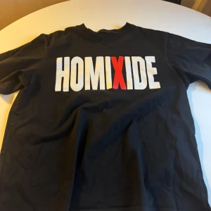 Svart HOMIXIDE t-shirt med tryck - Svart t-shirt med stort HOMIXIDE-tryck på bröstet i vitt och rött. Klassisk rund hals och korta ärmar. Skön och avslappnad passform, perfekt för dig som gillar streetwear och statement-plagg. 