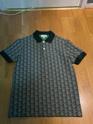 Mönstrad pikétröja från Lacoste - Säljer en svart och vit pikétröja från Lacoste med grafiskt mönster över hela plagget. Klassisk krage, två vita knappar framtill och den ikoniska krokodilloggan broderad på bröstet. Tröjan har korta ärmar och ribbade muddar.