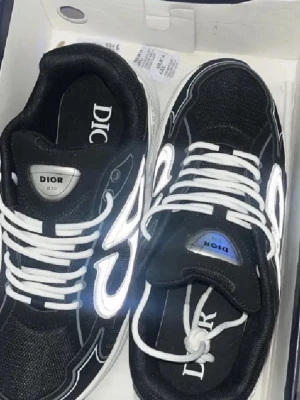 Svarta Dior B30 sneakers med vita detaljer - Säljer ett par svarta Dior B30 sneakers med vita och silverfärgade detaljer. Skorna har tydliga Dior-loggor på plösen och hälen, samt vita snören och en chunky vit sula. Materialet är en mix av mesh och syntet, vilket ger en sportig och modern vibe.