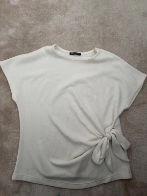 Vit topp med knyt från Zara - Säljer en vit topp från Zara i storlek M. Toppen har korta ärmar och en snygg knytning på sidan som ger en cool detalj. Materialet känns mjukt och har en lätt struktur.