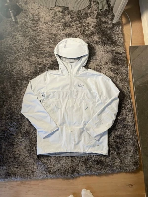 Vit vindjacka från Arc'teryx - Säljer en clean vit vindjacka från Arc'teryx med huva och dragkedja framtill. Jackan har två snedställda fickor med dragkedja och den ikoniska Arc'teryx-loggan på bröstet. Perfekt för dig som vill ha en stilren och funktionell jacka till vardags.