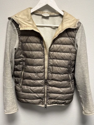 Beige och grå dunväst från Moncler - Snygg dunväst från Moncler i beige och grått med quiltad front och dragkedja. Västen har mjuka grå ärmar och insida i bomullsmix samt Moncler-logga på ärmen. Perfekt lager-på-lager-plagg med sportig känsla och två fickor med dragkedja. 100% äkta. Jackan är tillverkad innan 2022 så för att kunna se om den är äkta får man skanna qr koden som visas på bilden som sitter på insidan av jackan. Ny pris ca 9-10k .Skriv om ni har frågor!