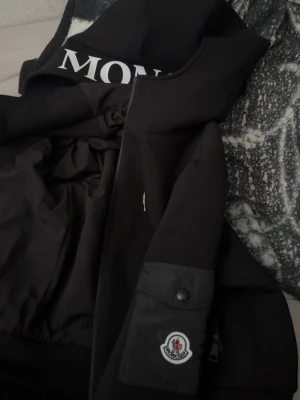 Svart Moncler vindjacka med huva - Svart kofta från Moncler med stor vit logga på huvan och klassisk Moncler-patch på ärmen. Jackan har dragkedja framtill, ficka med knapp och stilren design. Materialet är polyester och jackan har en loose passform som ger en chill vibe.