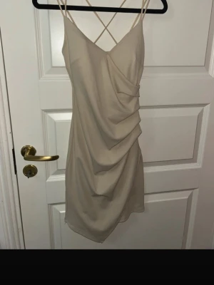 Beige ribbad kortklänning med omlott - Supersnygg beige kortklänning med tunna axelband som korsas i ryggen. Klänningen har ribbad struktur och omlottdetalj framtill som ger en draperad look. Perfekt för dig som gillar stilrena och trendiga plagg. Köpt utomlands 