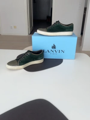 Lanvin Toe Cap Suede Low Sneakers - Snygga Lanvin sneakers i mörkgrön mocka med glansig svart tå och vita sulor. Skorna är nästintill helt perfekt skick Skorna har snörning och en klassisk låg profil. Lyxig känsla med detaljer i kalvskinn och gummisula. Perfekta för dig som vill ha något unikt och stilrent. Passar till allt och extremt Grisch