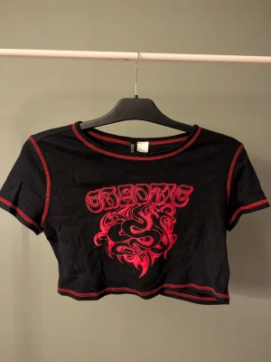 Svart croppad topp med röd orm - Cool svart croppad topp från Divided med röd kontrastsöm och tryck framtill med texten 'CHAOTIC' och en detaljerad orm. T-shirten har korta ärmar och är gjord i mjuk bomull. Perfekt för dig som gillar edgy streetstyle.