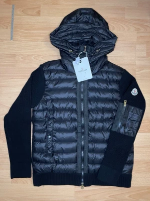 Svart Moncler pufferjacka med huva - Säljer en svart pufferjacka från Moncler med stickade ärmar och detaljer. Jackan har huva med snörning, dragkedja framtill och en ficka med dragkedja på ena ärmen. Klassisk Moncler-logga på ärmen och quiltad front i glansigt material. Liten i storlek!