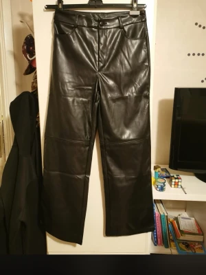 Svarta vida byxor i skinnimitation - Säljer ett par svarta byxor i skinnimitation med raka, vida ben och klassisk femficksdesign. Byxorna har hög midja och knapp- samt dragkedjestängning framtill. Perfekta för dig som vill ha en edgy look. Använd fåtal gånger. INTE äkta skinn. Välj helst insta box då både du och jag slipper frakt kostnaden.