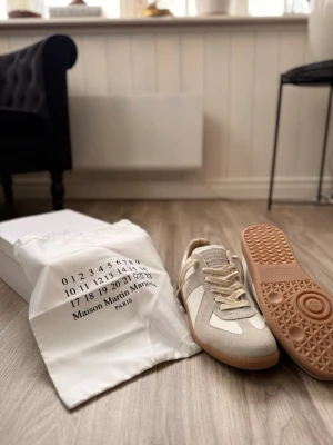 Maison Margiela sneakers i beige och vitt - Maison Margiela sneakers i beige mocka och vitt skinn med klassisk gummisula. Snygg retrostil med rund tå och snörning. Kommer med originalpåse och box. Perfekta för dig som gillar minimalistisk och clean design. Skriv våd frågor svarar snabbt! Pris kan diskuteras vid snabb afär men har en gräns 