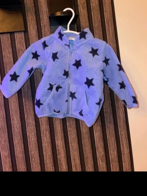 Blå fleecejacka med stjärnor storlek 68 - Mysig blå fleecejacka med svarta stjärnor och dragkedja framtill. Jackan har huva och två fickor, perfekt för kyliga dagar. Långärmad och i mjukt material som känns skönt mot huden. Storlek 68