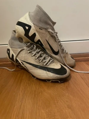 Nike fotbollsskor med högt skaft - Säljer ett par vita och svarta Nike fotbollsskor med högt skaft och snörning. Skorna har en modern design med stora Nike-loggor på sidorna och dobbar under sulan för bra grepp på planen. Perfekta för dig som vill sticka ut på fotbollsplanen.