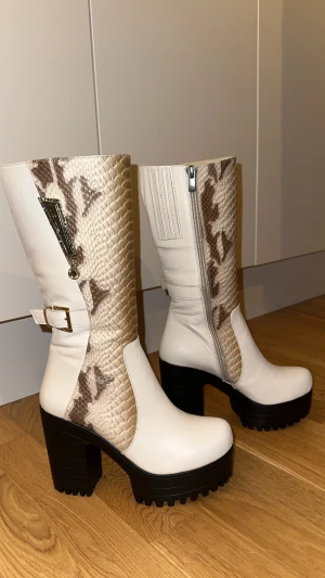 GUERO Stövlar, vit, storlek 36 - äktaskinn. I nytt skick  - 100 % äktaskinn, vita stövlar med ormskinnsmönster och dragkedja. Stövlarna har en hög klack, väldigt bekväma och varma. Dekorativ-guldiga detaljer med text på sidan.