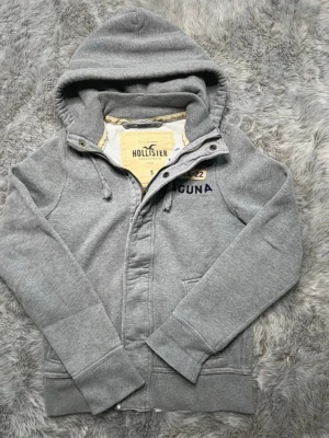 Hollister tröja  - Grå hoodie från Hollister i mjukt och bekvämt material. Klassisk modell med huva och knäppning framtill. Liten Hollister-logga/tryck på bröstet. Perfekt som vardagströja eller lager på lager.