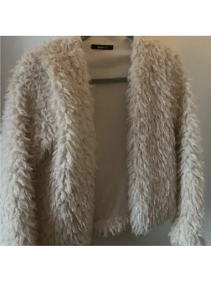 Beige fluffig pälskofta från Ginatricot - Supermjuk och fluffig beige pälskofta från Ginatricot. Koftan har lång ärm, öppen framtill och är tillverkad i ett syntetiskt pälsmaterial som ger en cool och trendig look. Perfekt för dig som vill sticka ut och hålla dig varm under kyliga dagar.