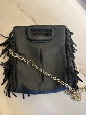 Svart axelväska med fransar från Maje - Säljer en svart axelväska från Maje i skinn med fransar på sidorna och en chunky guldfärgad kedja. Väskan har ett stilrent handtag upptill och minimalistisk design med coola detaljer. Köpte den på Maje i Stockholm men har inget kvitto kvar. Nypris 3145 kr och säljer den nu för 490 kr. 