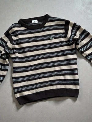 Vitage lacosta wool striped sweater - Säljer en riktigt snygg stickad tröja från Lacoste med breda svarta och beige ränder samt tunna grå detaljer. Klassisk rund halsringning och ikoniska krokodilloggan på bröstet. Perfekt för dig som gillar retrovibbar och vill ha något både stilrent och chill.