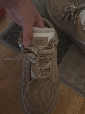 Beige sneakers från UGG med snörning - Säljer ett par snygga beige sneakers från UGG med breda snören och mysigt foder. Skorna har detaljer i mocka och mesh samt en rund tå. Perfekta för dig som vill ha både stil och komfort under höst och vinter.