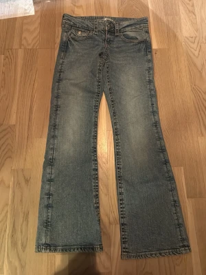 Blåa gina tricot  jeans  - Snygga blå jeans med bootcut passform och klassisk femficksdesign. Jeansen har dekorativa knappar på bakfickorna och en lätt tvättad look. Tillverkade i jeansmaterial med normal passform och låg midja.