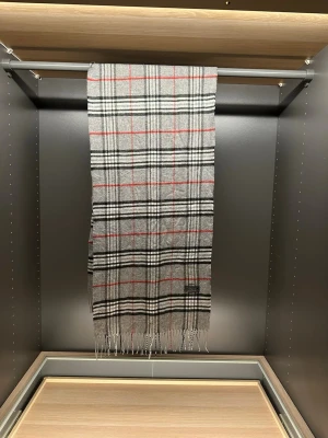 Grå rutig halsduk från Burberrys - Klassisk grå halsduk från Burberrys i 100% lammull. Rutigt mönster i svart, vitt och rött med fransar i kanterna. Tidlös accessoar som ger en snygg touch till din outfit. Tillverkad i England.