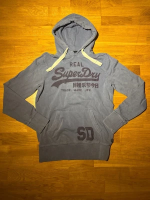 Mörkblå Superdry hoodie - Säljer en snygg mörkblå superdry hoodie en mörkare blå text med vintage look. Se bild 2 för mer exakt representation av färgen. Inga defekter, en slim passform och passar även S lika väl som M. Hör av dig vid frågor eller annat…