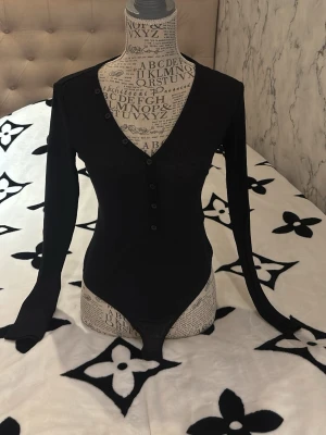 Svart långärmad body med knappar - Säljer en svart långärmad body med v-ringning och knappar framtill. Tight passform och mjukt stretchigt material som sitter snyggt på kroppen. Perfekt att styla med jeans eller kjol för en clean look.