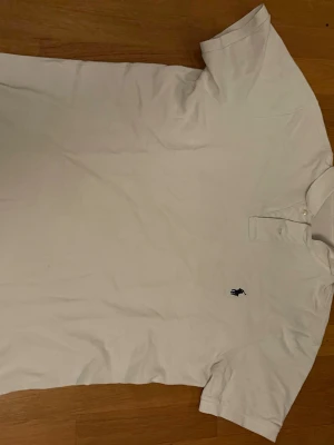 Vit pikétröja från Ralph Lauren - Klassisk vit pikétröja från Ralph Lauren med korta ärmar och den ikoniska broderade loggan på bröstet. Tröjan har krage och knappar vid halsen. Tillverkad i mjuk bomull som känns skön mot huden.