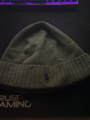 Grå mössa från Polo Ralph Lauren - Säljer en stilren grå mössa från Polo Ralph Lauren i 100% merinoull. Mössan har ribbad kant och klassisk broderad logga framtill. Perfekt accessoar för kyliga dagar och enkel att matcha med olika outfits. Säljes pga att den inte används längre.