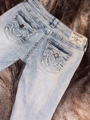 True Religion Joey Low Rise Flare Jeans - Säljer ett par ljusblå True Religion Joey jeans med låg midja och utsvängda ben. Jeansen har snygga detaljerade bakfickor med knappar och tydliga sömmar. Klassisk tvättad denim-look och cool etikett bak i midjan.