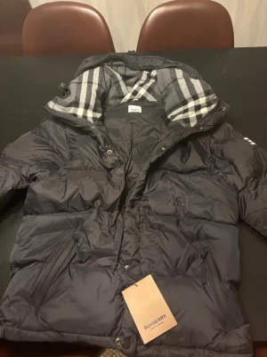 Svart pufferjacka från Burberry - Säljer en svart pufferjacka från Burberry med klassiskt rutigt foder.  Perfekt för kalla dagar och riktigt stilren. Säljer den här jackan då den tyvärr inte passar mig. Jackan är sprillans ny.