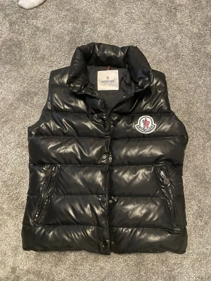 Svart dunväst från Moncler - Säljer en svart dunväst från Moncler med hög krage och ikonisk logga på bröstet. Västen har blanka paneler, dragkedja framtill och två fickor med dragkedja. Perfekt för lager-på-lager och riktigt snygg till streetwear. Har inget äktahetsbrvis tyvärr men jag är nästan 100% på att den är äkta.