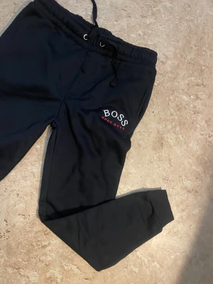 Svarta mjukisbyxor från Hugo Boss - Svarta mjukisbyxor från Hugo Boss med broderad logga på vänster ben. Byxorna har resår och snörning i midjan samt muddar vid bensluten. Tillverkade i mjukt bomullsmaterial för maximal komfort. Perfekta för chill dagar eller när du vill ha en clean look.