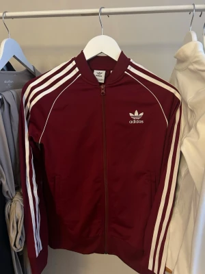 Vinröd Adidas ziptröja - Säljer en vinröd Adidas ziptröja med klassiska vita ränder längs ärmarna och Adidas-logga på bröstet. Tröjan har lång ärm, ribbade muddar och dragkedja framtill. Perfekt för en sportig och avslappnad look.
