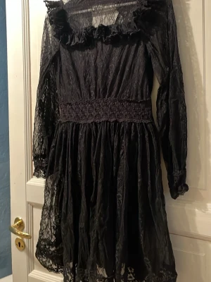 BY MALINA svartspets klänning - Supersnygg svart kortklänning i spets med långa ärmar och volangdetaljer vid halsringningen. Klänningen har markerad midja med broderade detaljer och transparenta partier på ärmar och nederkant. Perfekt för dig som vill ha en romantisk och edgy look.