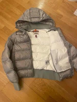 Grå Parajumpers pufferjacka med huva - Säljer en grå pufferjacka från Parajumpers, modell Sheen. Jackan har huva, ribbade muddar och dragkedja framtill. Insidan är vit och jackan har en klassisk patch på ärmen. Perfekt för kalla dagar och riktigt snygg till streetwear.