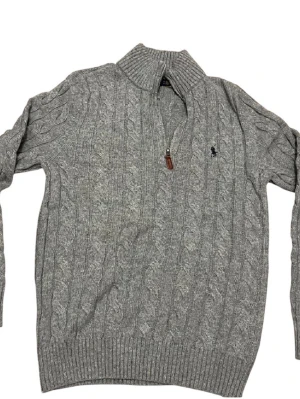 Grå stickad tröja Polo Ralph Lauren - Snygg grå stickad tröja från Polo Ralph Lauren med klassiskt kabelmönster och halv dragkedja vid kragen. Tröjan har långärmade ribbade muddar och en broderad marinblå logga på bröstet. Perfekt för dig som gillar stilren och tidlös design.