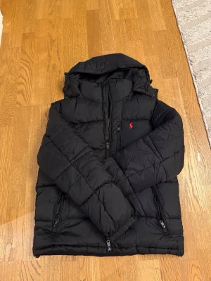 Svart pufferjacka från Polo Ralph Lauren - Säljer en svart pufferjacka från Polo Ralph Lauren med huva och dragkedjor. Jackan har det klassiska röda Polo-logot på bröstet och flera praktiska fickor med dragkedja. Perfekt för kalla dagar och väldigt stilig. Aldrig använd.