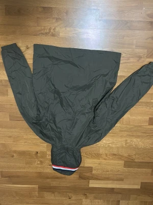 Svart Moncler vindjacka med huva - Säljer en oliv grön vindjacka från Moncler med huva och dragkedja framtill. Jackan har Moncler-logga på ärmen, två fickor med knappar och snygg detalj med röd, vit och blå rand på huvan. Tillverkad i lätt polyester, perfekt för blåsiga dagar. Storlek 4 som är ungefär L. 