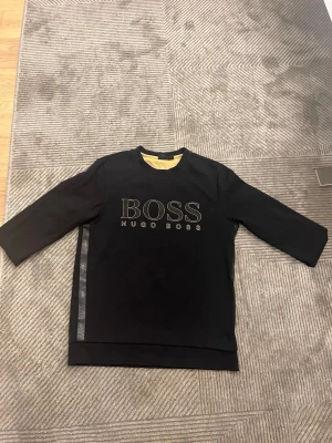 Svart sweatshirt från Hugo Boss M - Säljer en svart sweatshirt från Hugo Boss i storlek M. Tröjan har ett stort BOSS-tryck framtill och diskreta banddetaljer längs sidan. Klassisk rund halsringning och trekvartsärm ger en clean och modern look. Som passar folk med storlek S och möjligen folk med storlek xs