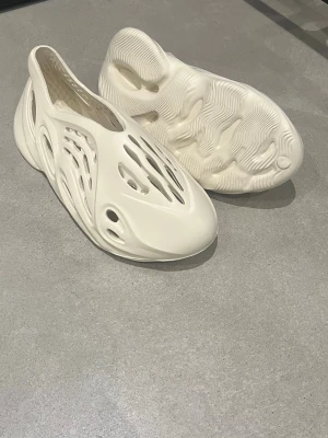 Adidas Yeezy Foam Runner vit - Säljer ett par Adidas Yeezy Foam Runner i helvit färg. Skorna har futuristisk design med många ventilationshål och är tillverkade i lätt syntetmaterial. Perfekta för dig som vill ha något unikt och bekvämt på fötterna.