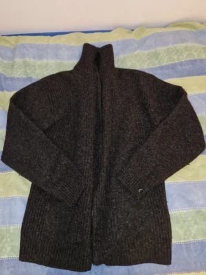 Mörkgrå stickad kofta från KappAhl - Mysig mörkgrå stickad kofta från KappAhl i storlek M. Koftan har hög krage, långa ärmar och ribbade muddar. Perfekt för kyliga dagar och enkel att matcha med olika outfits.